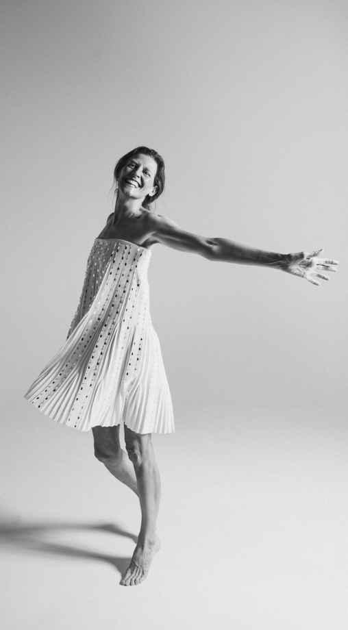 Mujer girando con un vestido blanco, brazo extendido, descalza sobre fondo blanco, sonriendo.