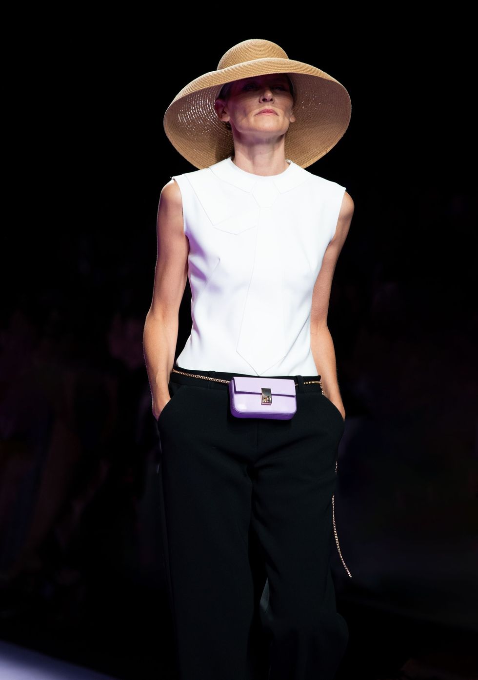 Mujer con blusa blanca y pantalones negros, sombrero de paja, caminando por una pasarela, fondo negro.