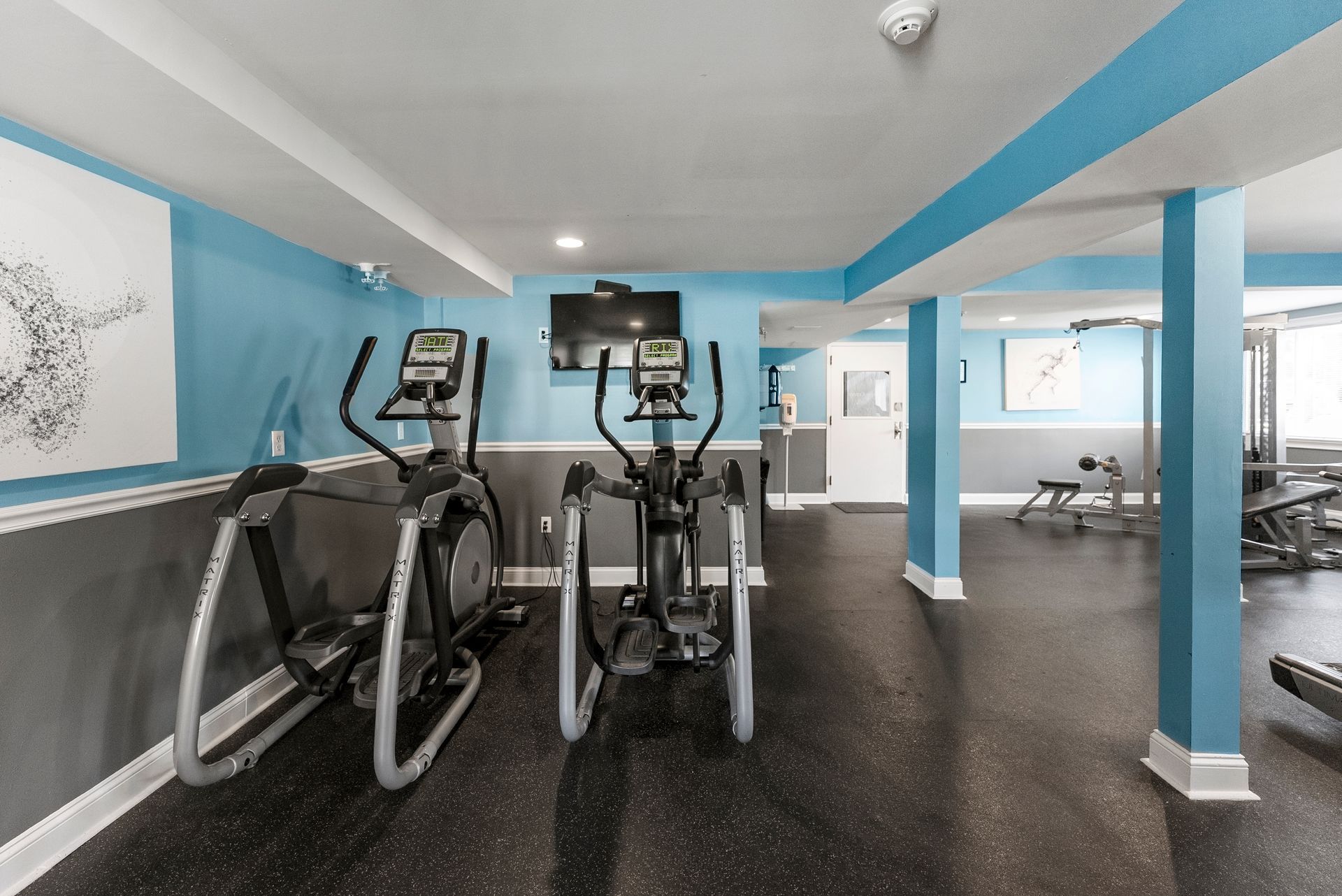 Gym with elliptical machines, TV, and weight machines; blue, gray, and white walls; dark floor at The Groves at Milford in Milford, MA.