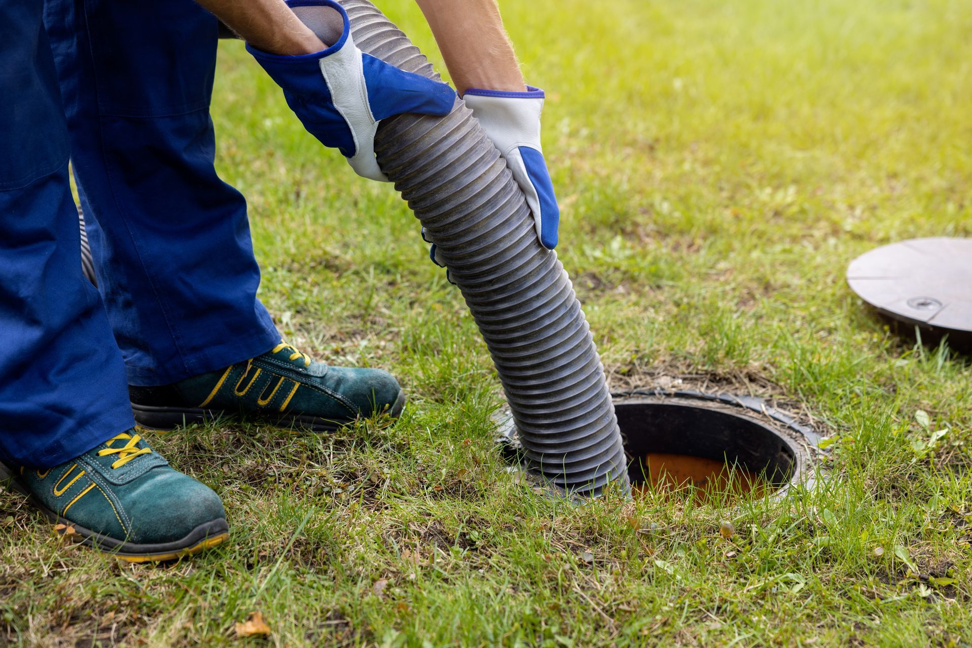 Person in blue overalls and gloves inserting a hose into a septic tank in a grassy yard.