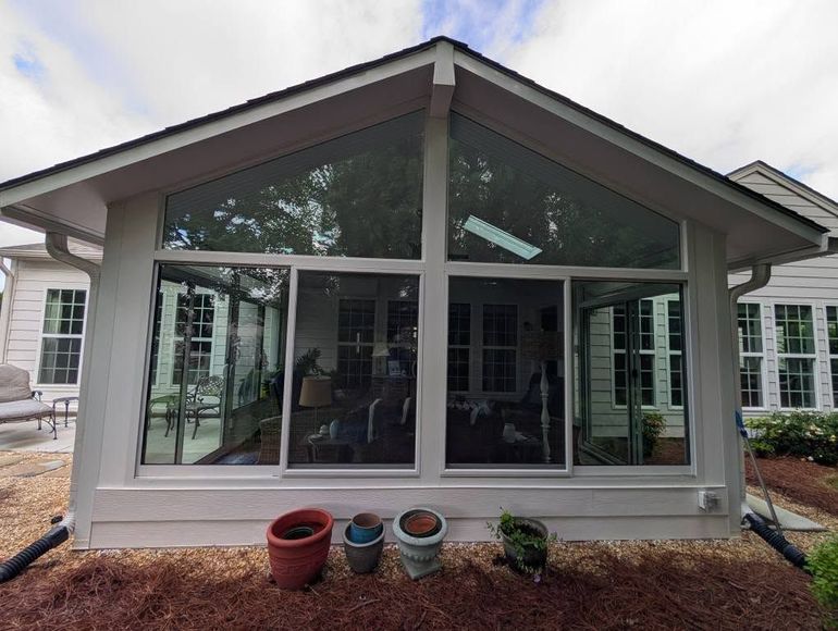 Sunroom with large windows, white trim, and a gabled roof; several potted plants sit below the windows.