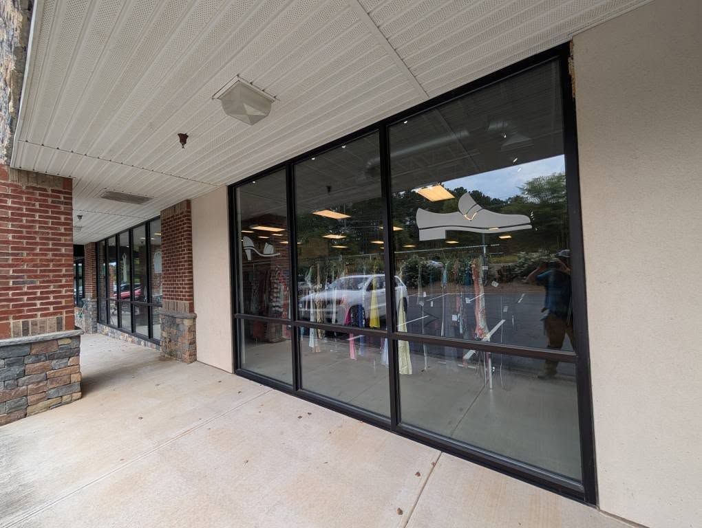 Exterior of a store with large windows, reflecting outdoor scene and shoe display; sidewalk and awning overhead.