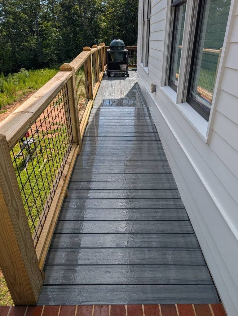 A narrow gray deck with a metal railing and a grill, next to a white house with a grassy background.