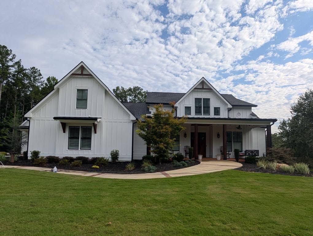 White farmhouse with black accents, front porch, and landscaped yard under a cloudy sky.
