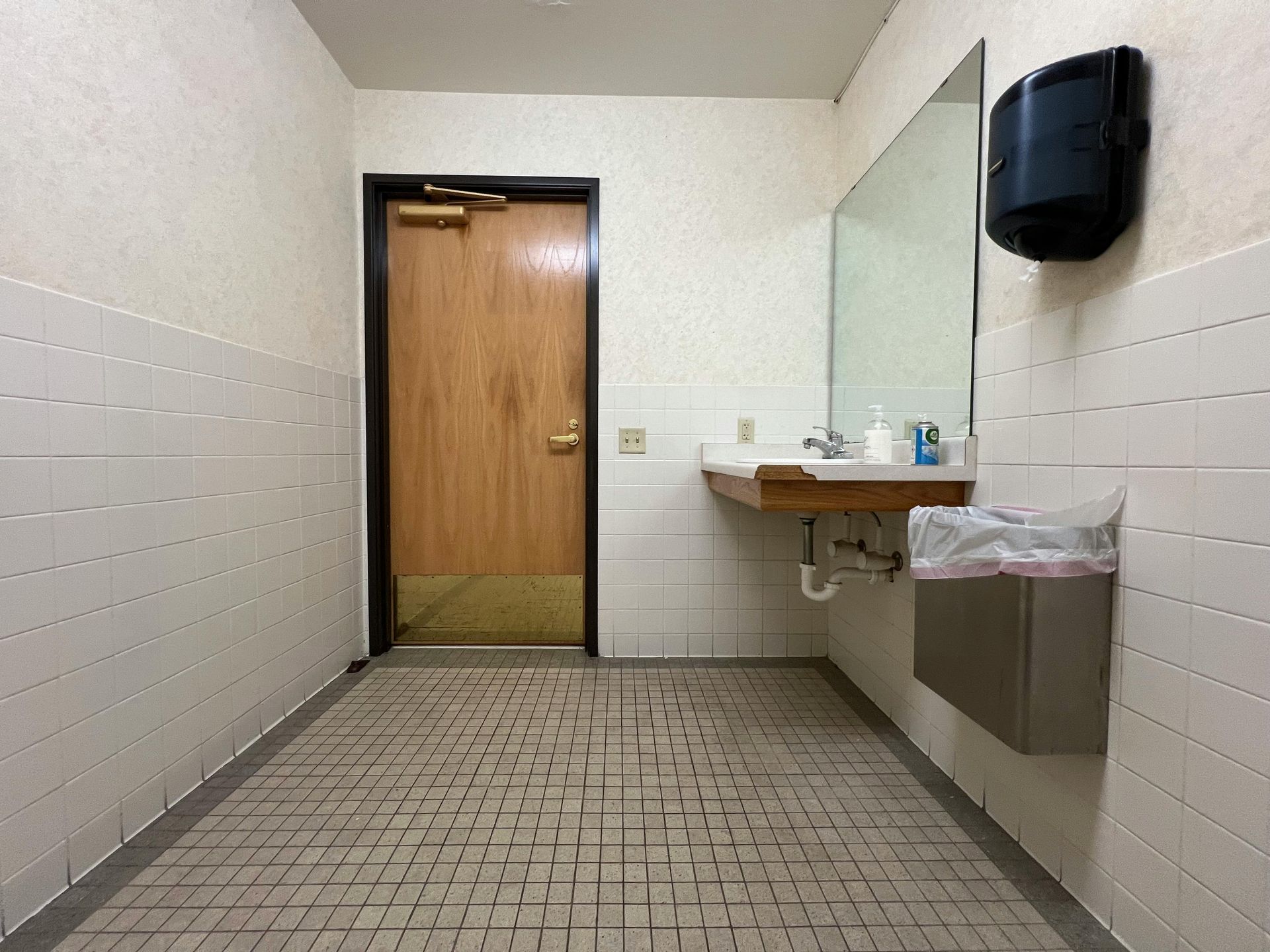 Public restroom with white tiled walls and floor. Sink, mirror, door and trash bin are visible.