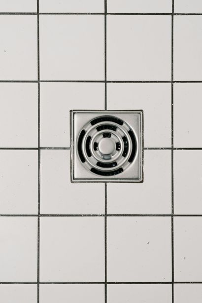 Square metal shower drain in white tiled floor.