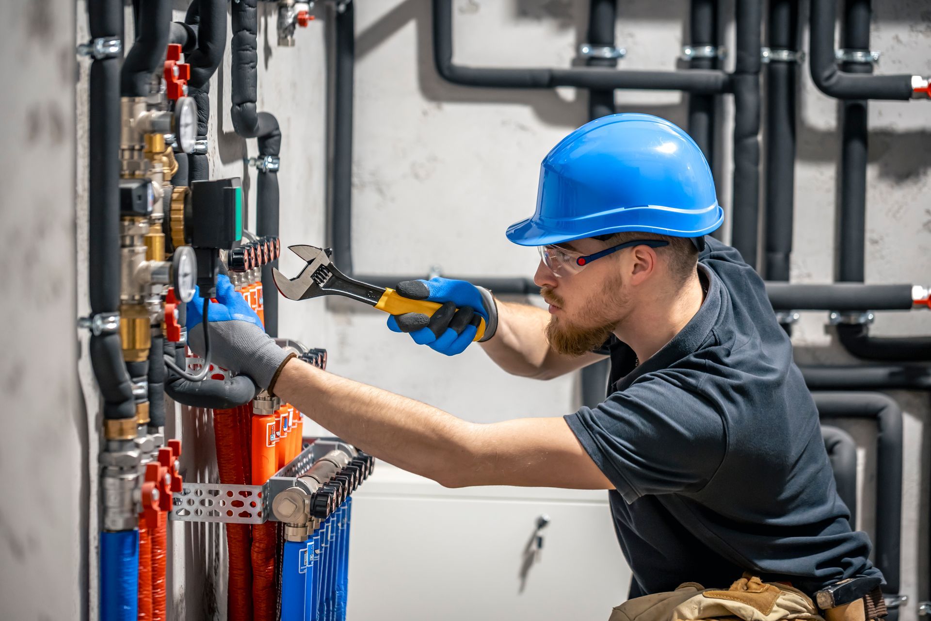 Plumber in a blue hard hat using a wrench on pipes, in an industrial setting.