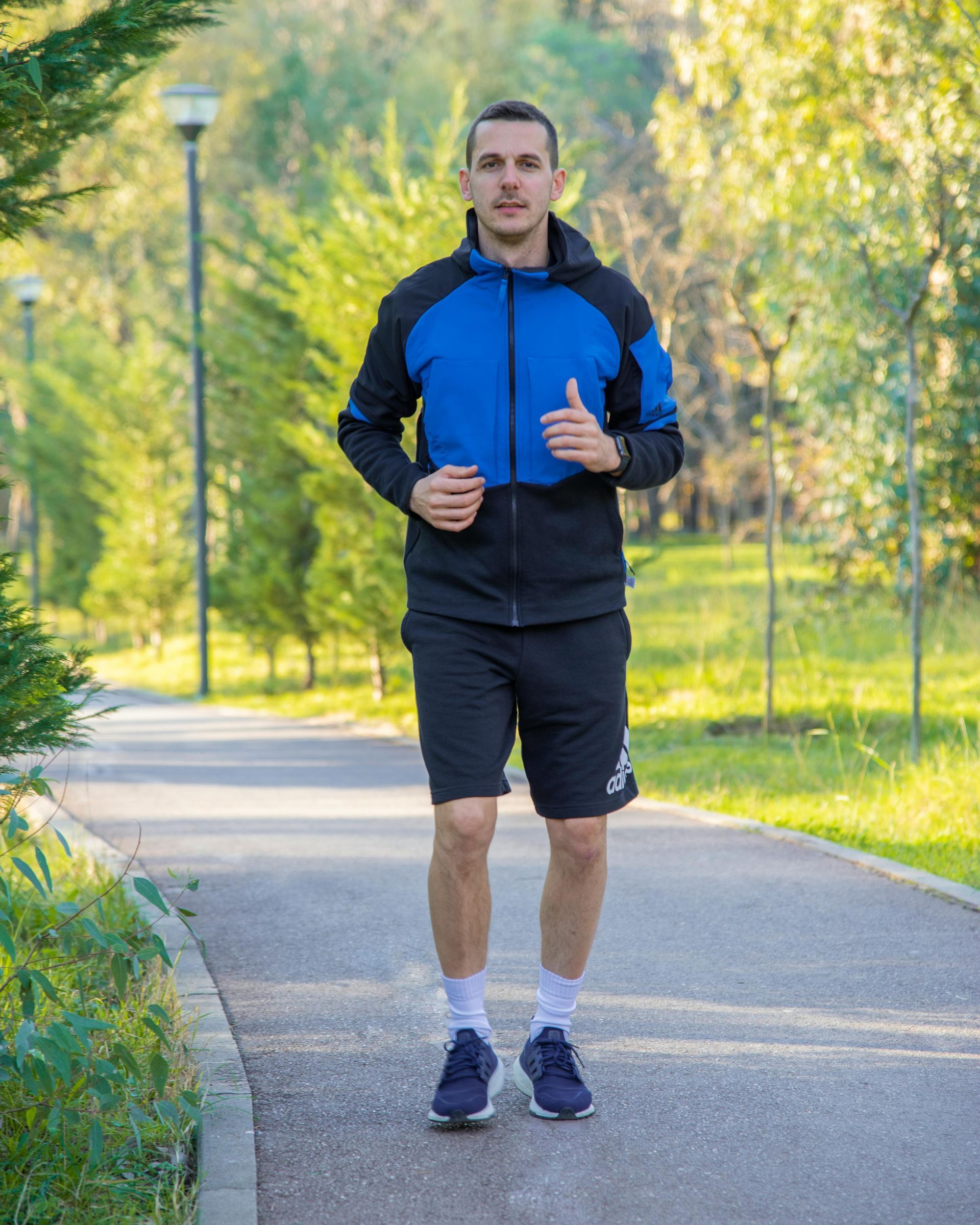 A man in a blue jacket and black shorts is running down a path in a park.