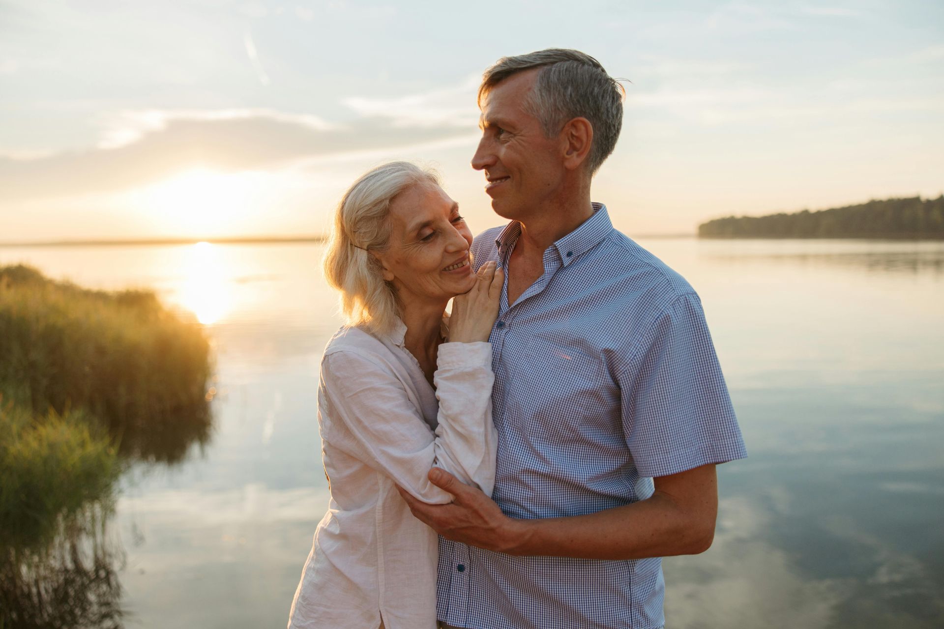 An elderly couple is hugging each other on the shore of a lake at sunset.