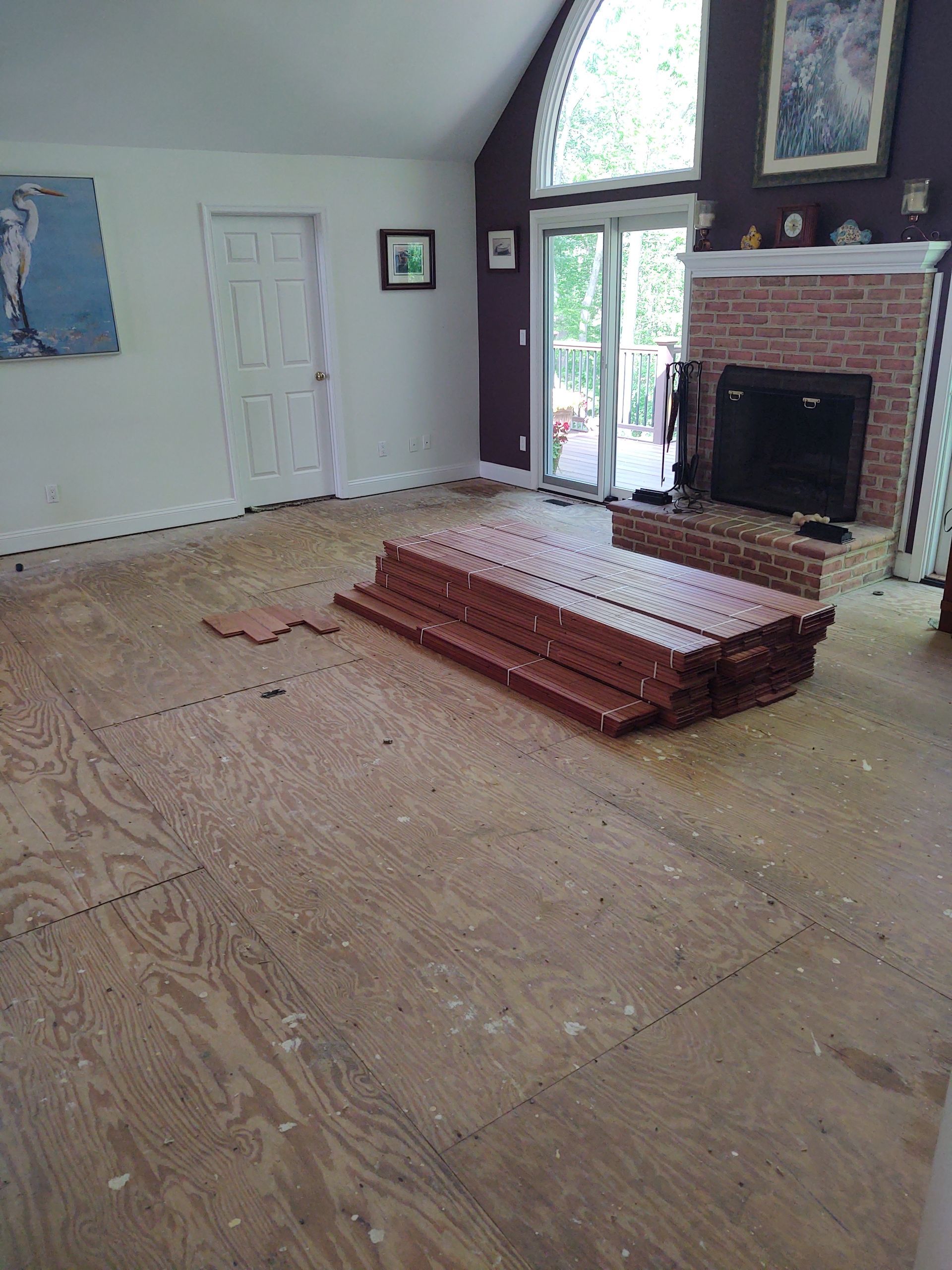 Interior of a room with plywood subfloor and stacked flooring materials; fireplace and doorway visible.