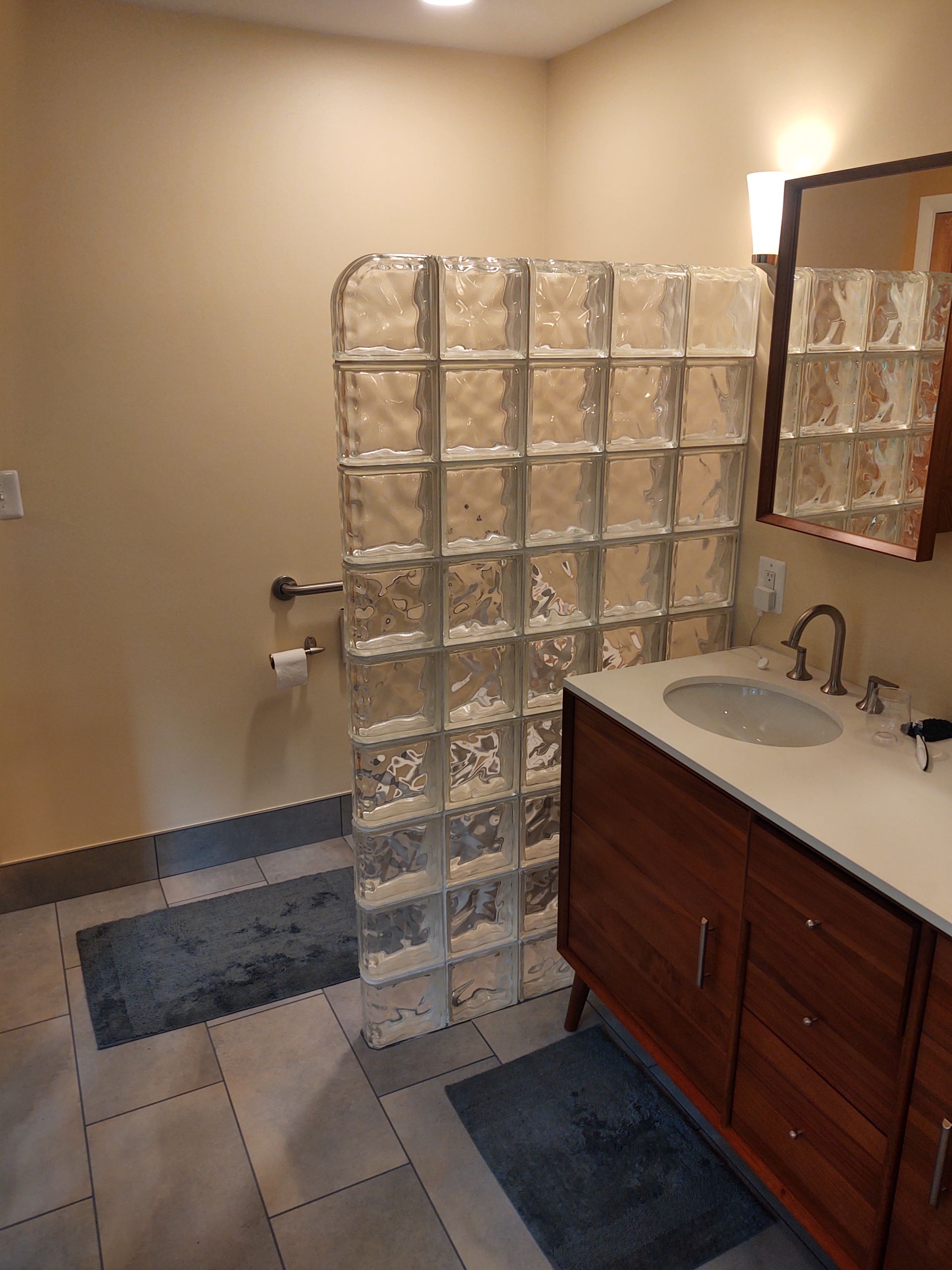 Bathroom with glass block shower wall, vanity, and toilet. Beige walls and blue rugs.