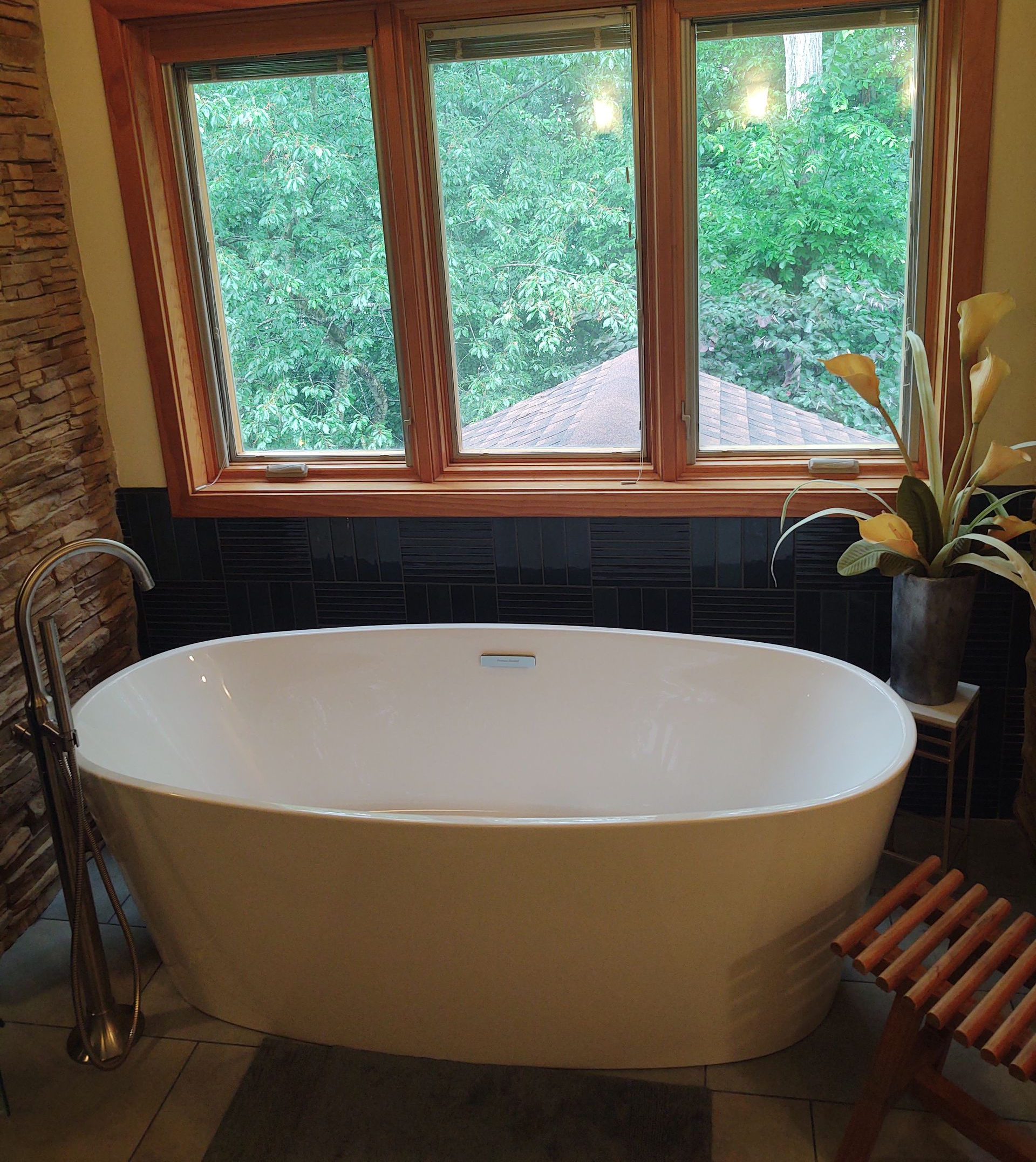 Freestanding white bathtub under a window overlooking a lush green canopy.