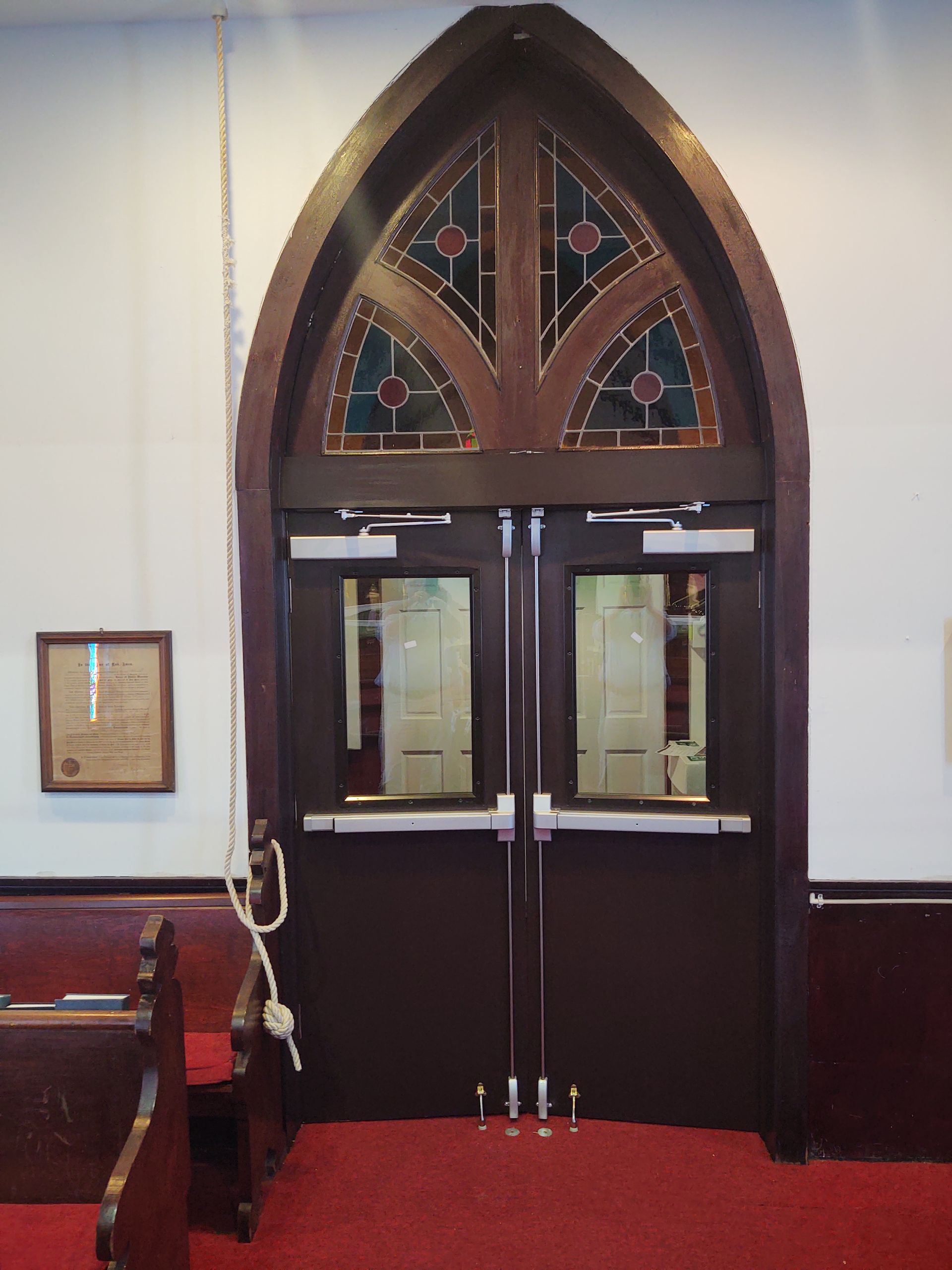 Church doors with arched stained glass window above, pull-down rope. Red carpet, wooden pews.