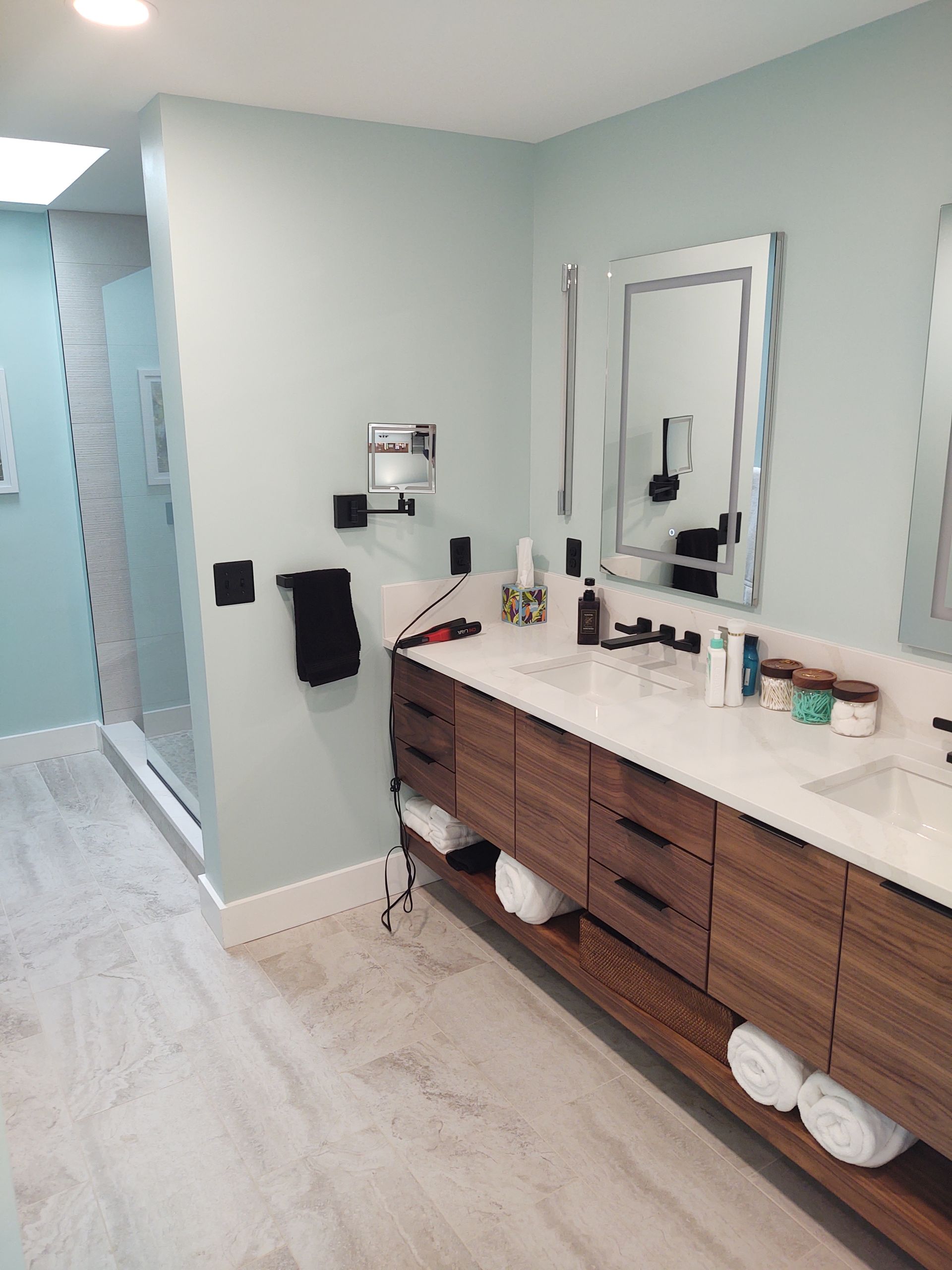 Spacious bathroom with light blue walls, wood vanity, two sinks, large mirrors, and a glass shower.