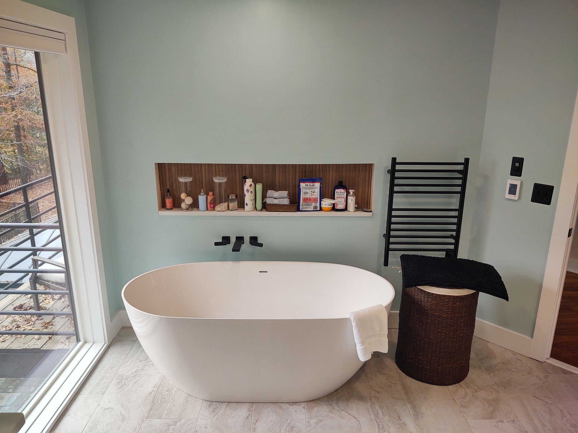 A modern bathroom with a white tub, light blue walls, built-in shelf with toiletries, and a black towel rack.