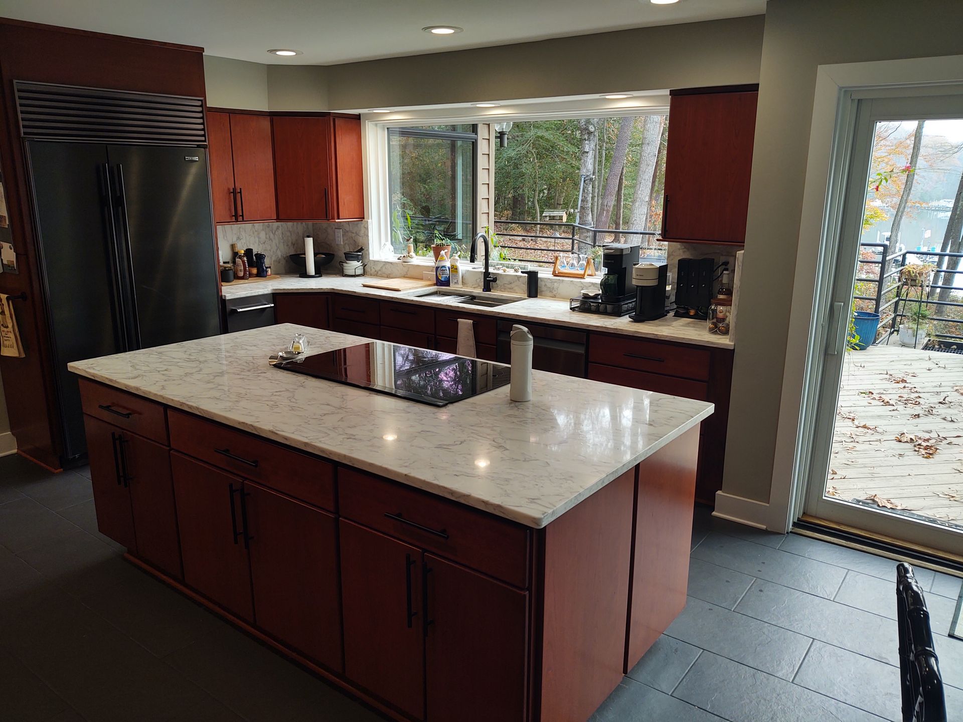 Kitchen with island, dark cabinets, white countertops, and stainless steel appliances. Large window overlooks trees.