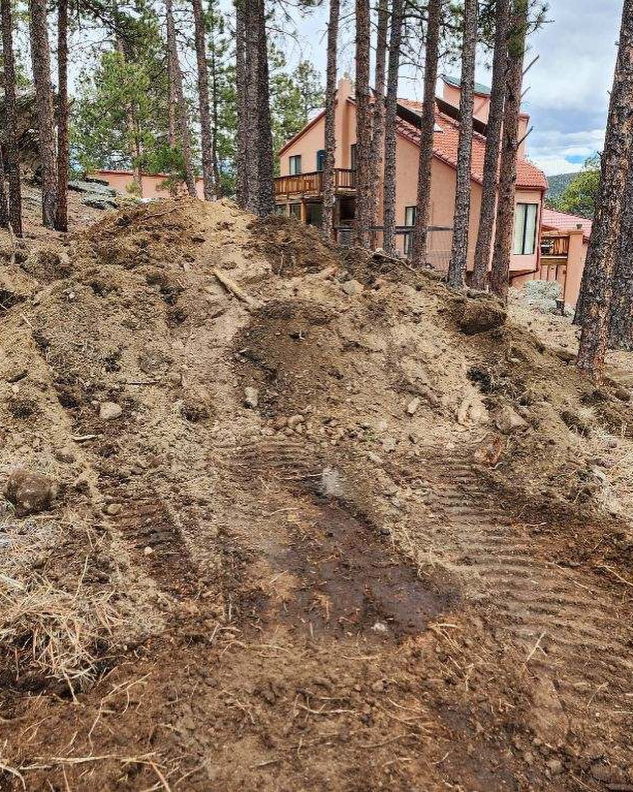Earth-moving equipment tracks on a hillside, with a house visible in the background, surrounded by trees.