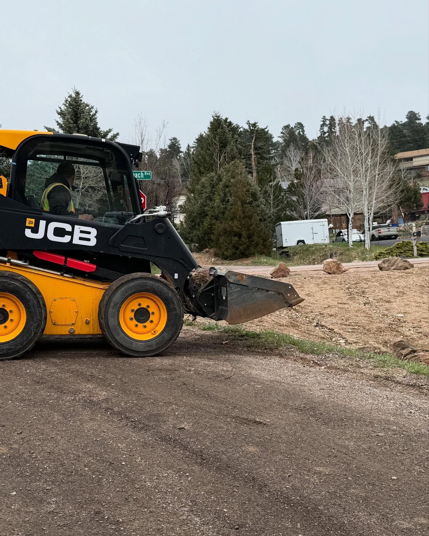 Yellow and black JCB skid steer on a construction site with trees and a white trailer in the background.