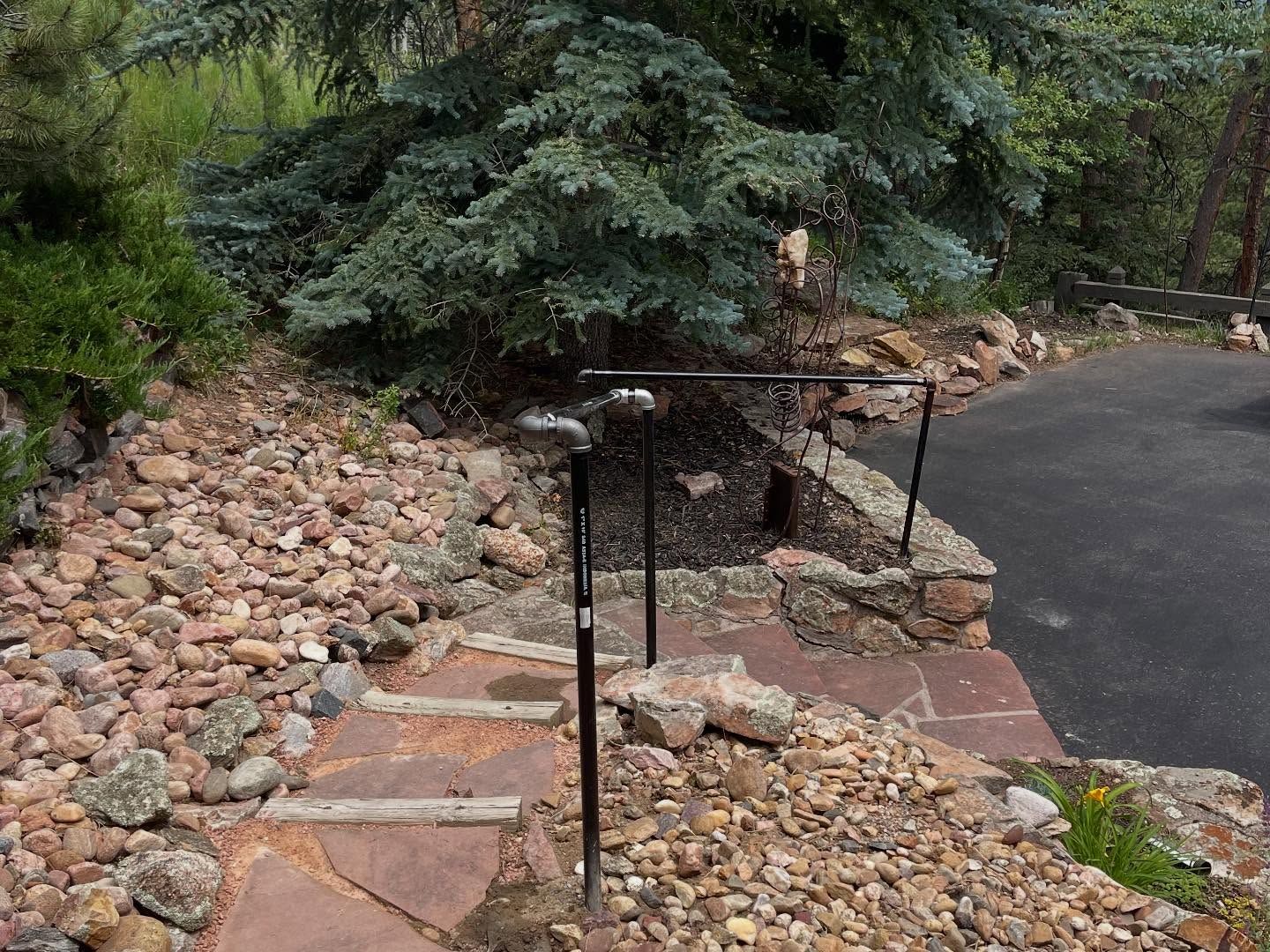 Stone steps with a black railing lead up from a driveway, surrounded by rocks and a blue spruce tree.