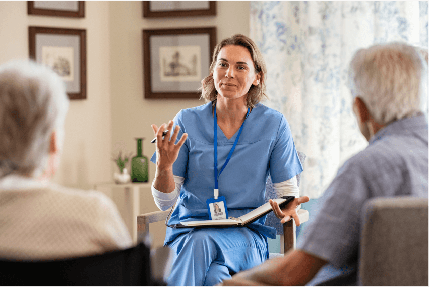 Healthcare worker in blue talks to an elderly couple in a homey setting