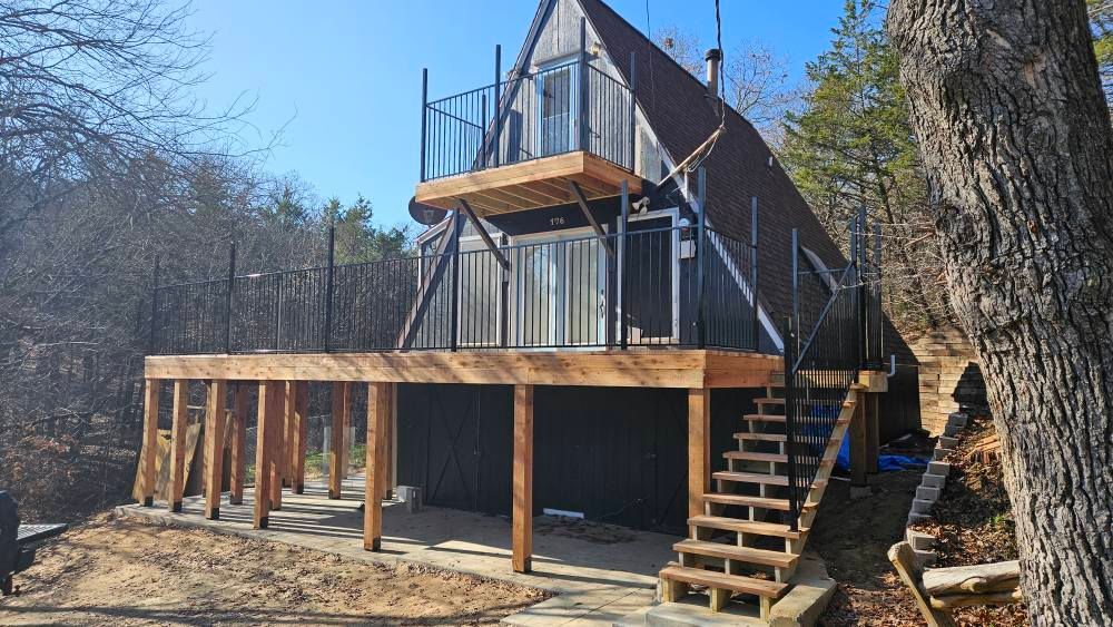 A small house with a large deck and stairs is surrounded by trees.