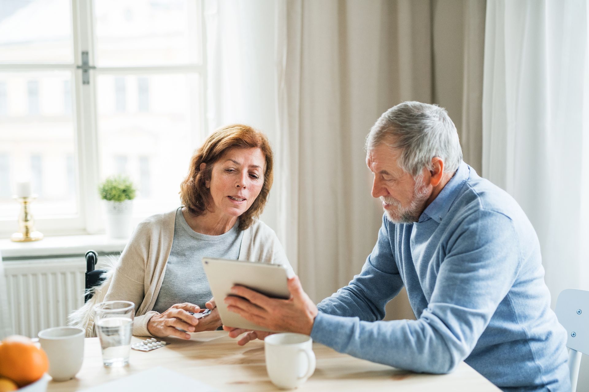 An elderly couple is sitting at a table looking at a tablet.