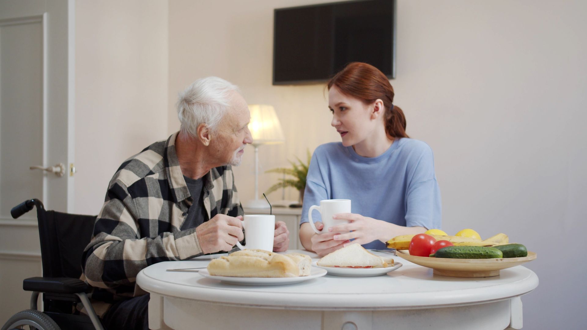An elderly man in a wheelchair is sitting at a table with a nurse.