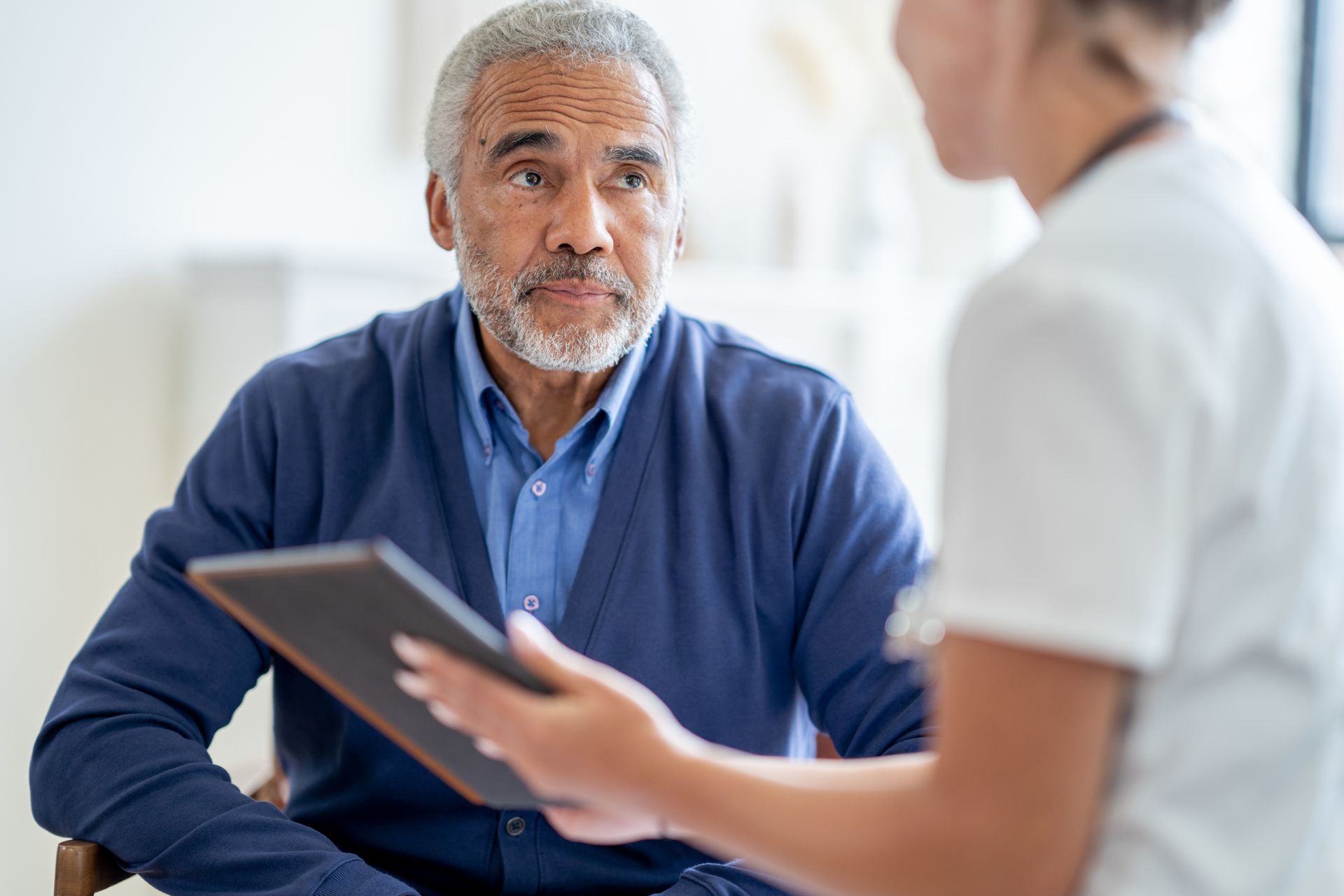 An elderly man is sitting in a chair talking to a nurse while holding a tablet.