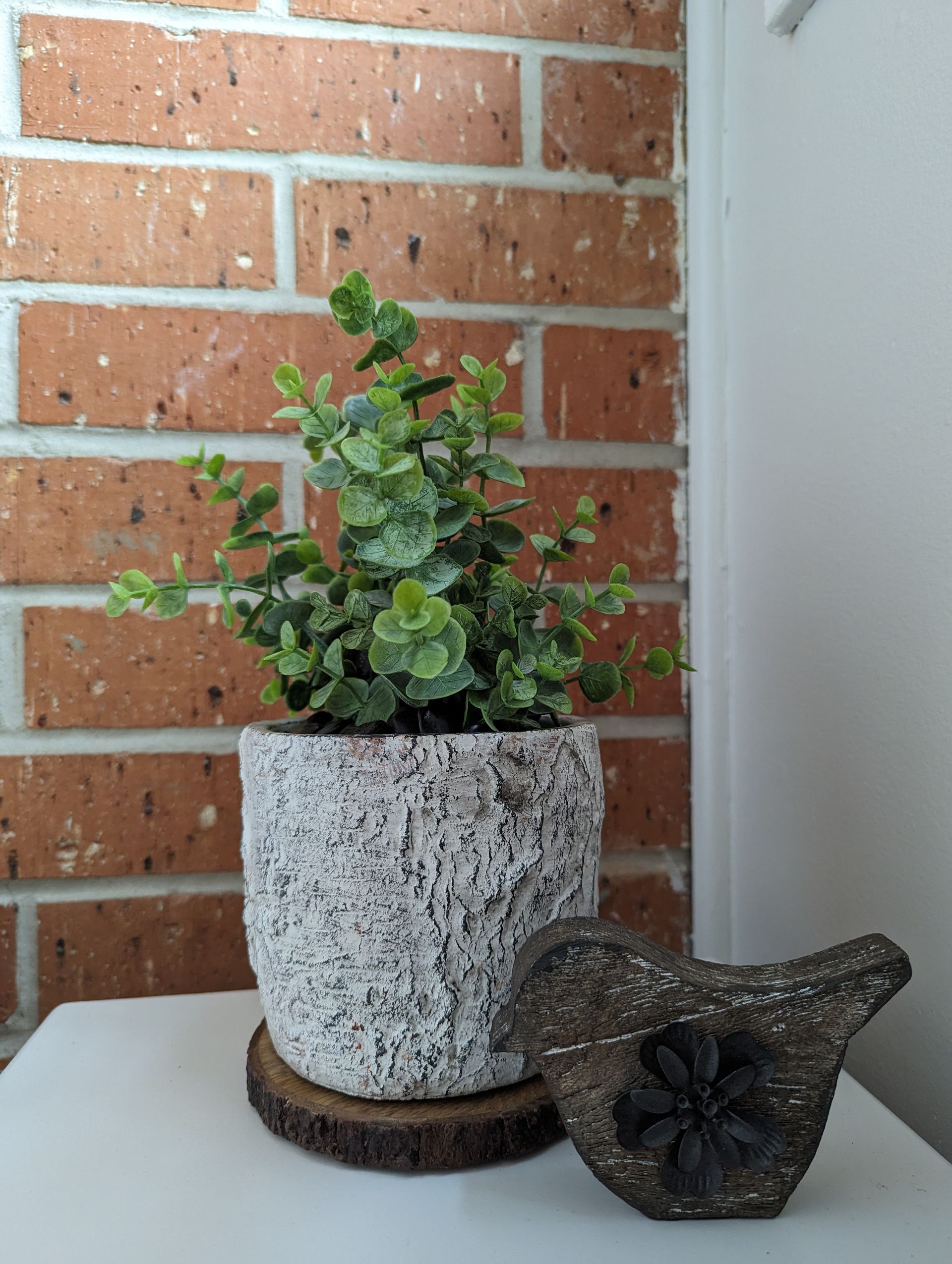 A potted plant and a bird figurine are sitting on a table in front of a brick wall.