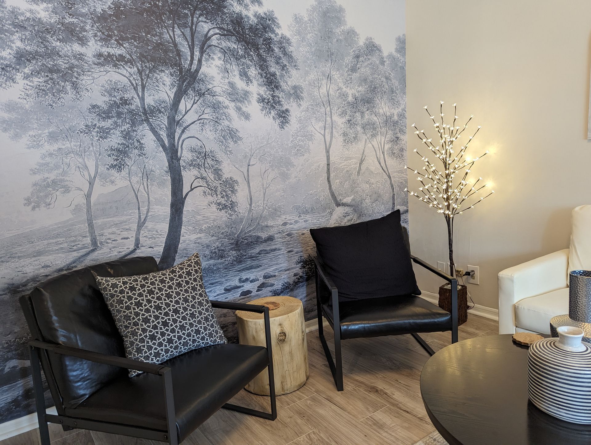 A living room with two chairs , a table , and a painting on the wall.