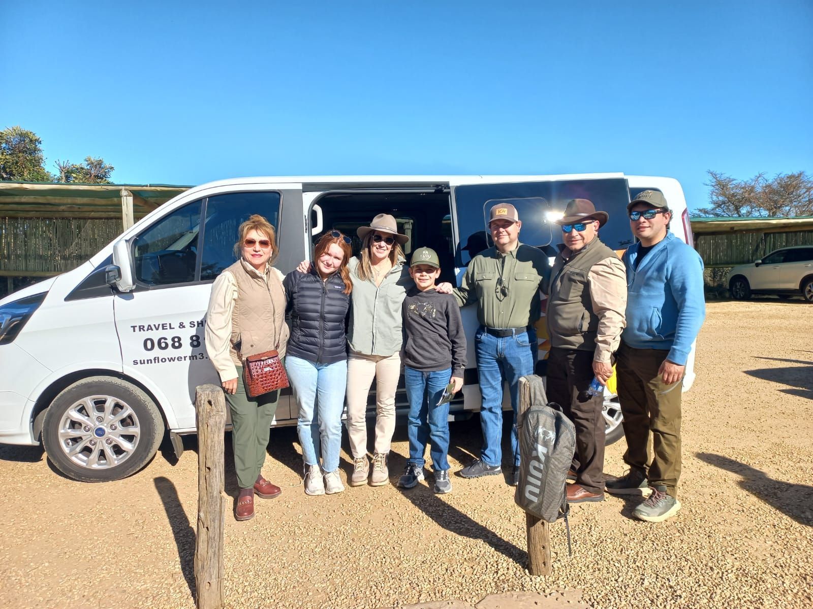 Group of people stand in front of a white van with open door in a sunny outdoor setting.