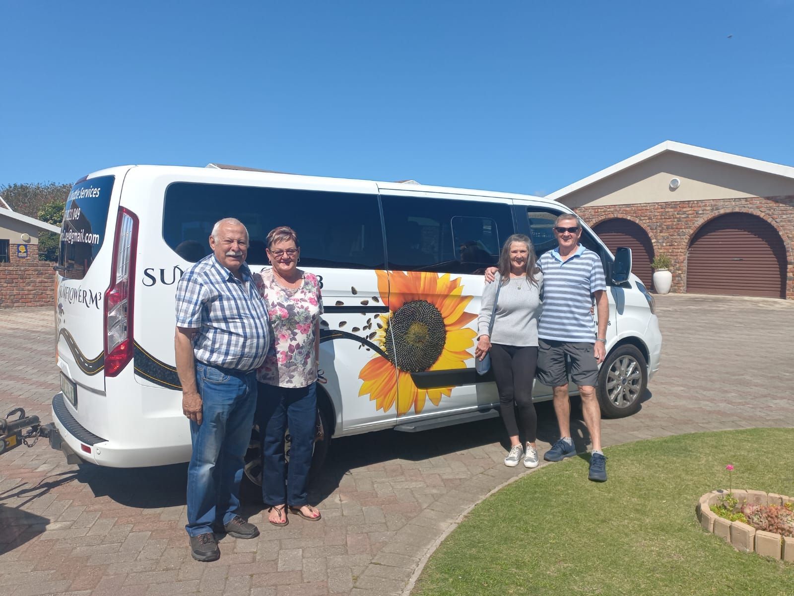 Four people stand in front of a white van with a sunflower graphic on the side, under a clear blue sky.