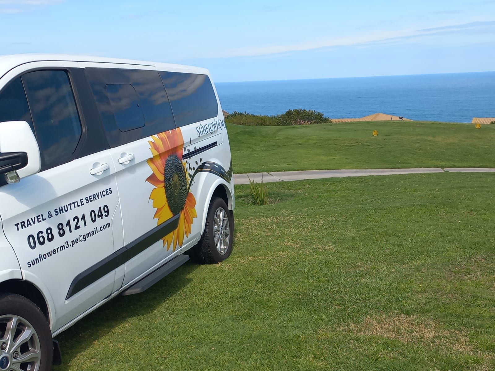 White van with sunflower logo parked on green grass, ocean in the background.