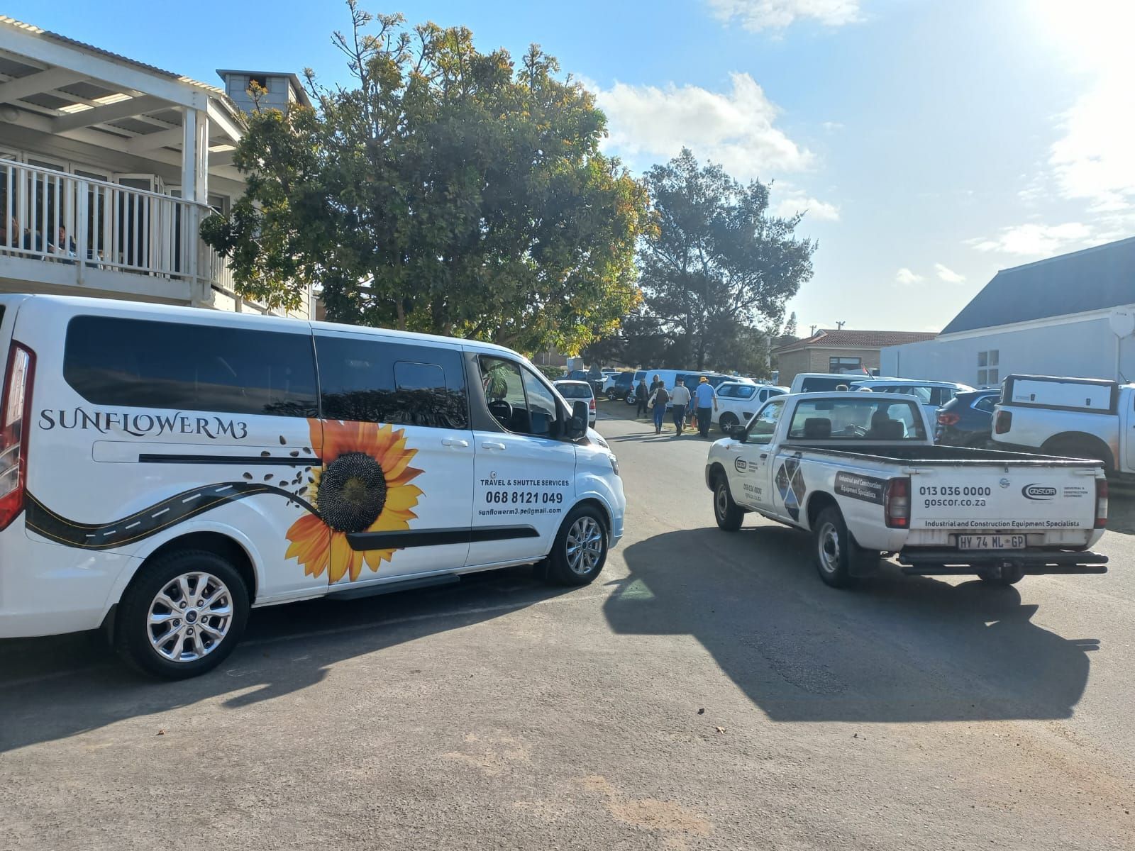 A white van and truck, both with logos, parked outside a building. People stand in the background.