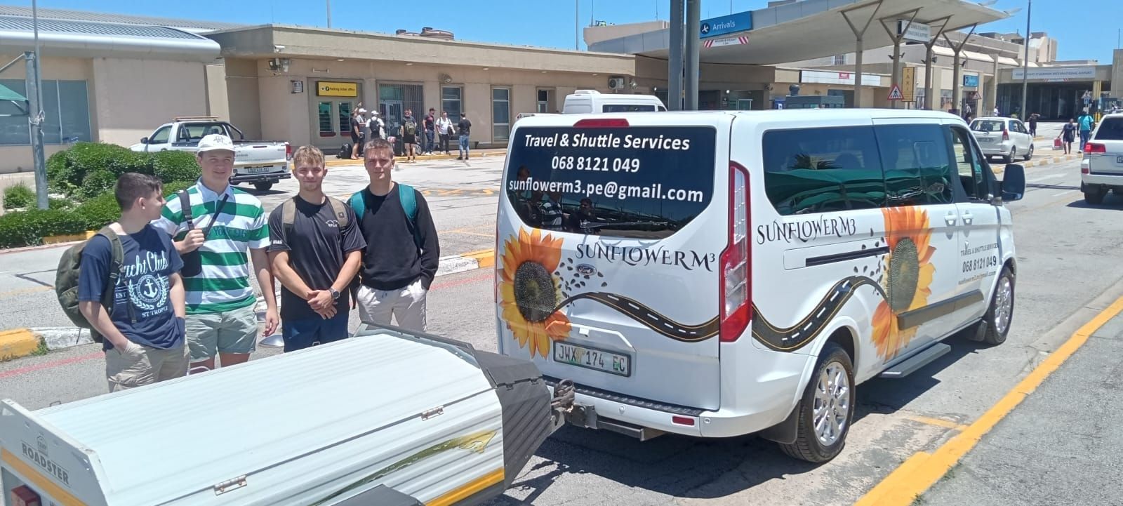 Four people stand near a white van decorated with sunflowers at an airport.