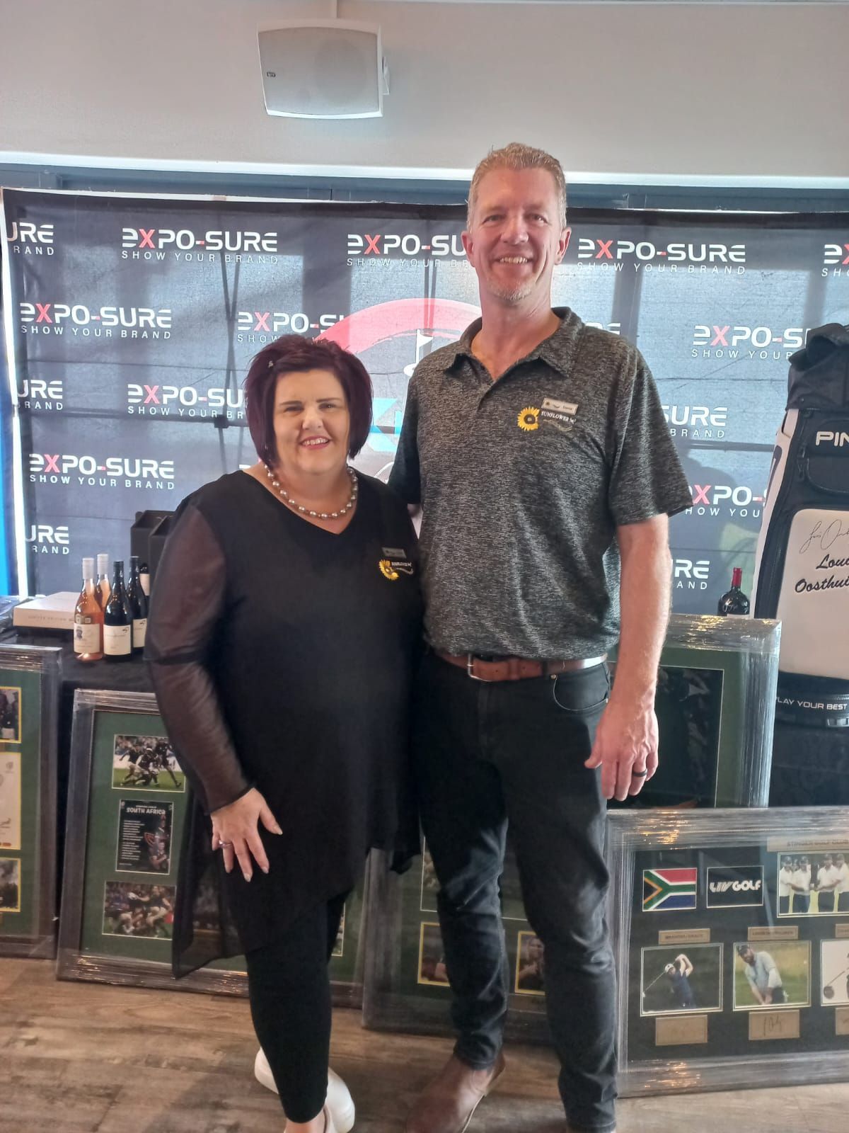 Two people stand in front of a backdrop with logos. A woman in black and a man in grey shirt pose for a photo.