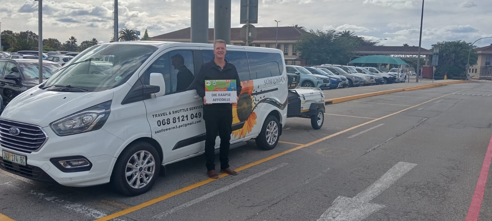 Man stands next to a white van holding a sign in a parking lot. Other cars and buildings are in the background.