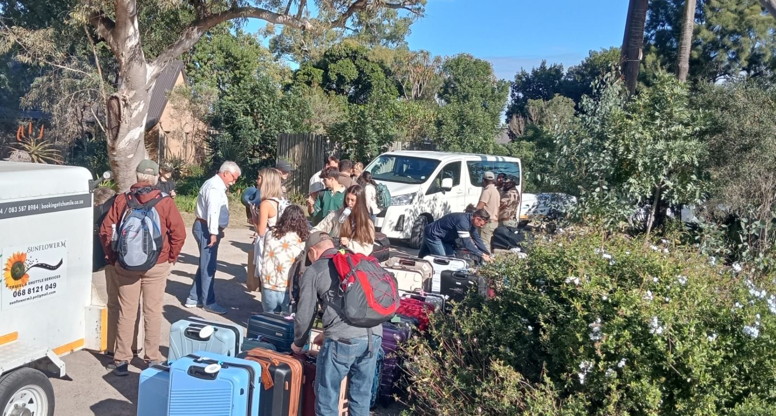 People unloading luggage from a van and trailer in a sunny, tree-lined area.