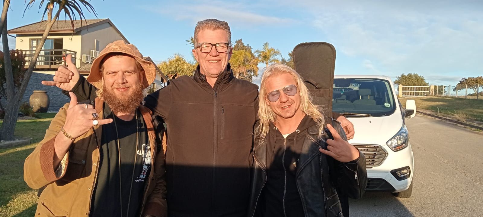 Three people posing outdoors next to a white van, with a building in the background. Two are giving hand gestures.
