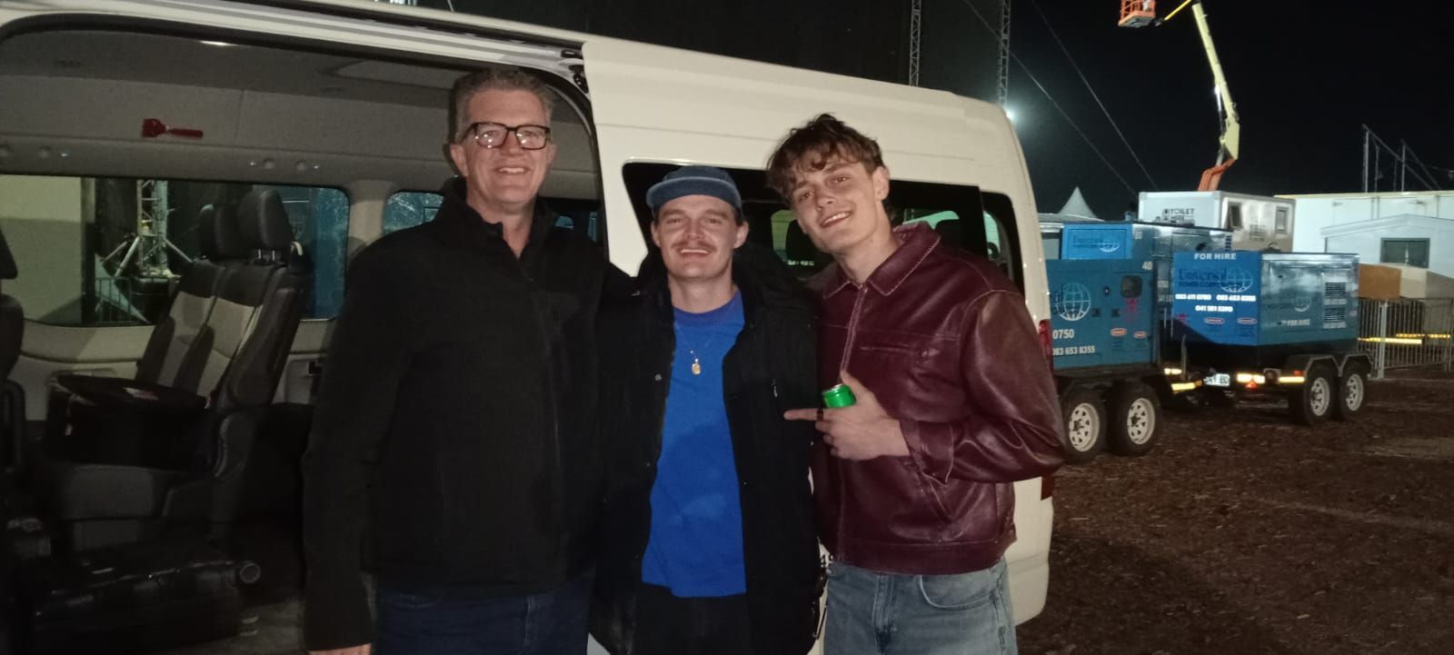 Three people pose for a photo in front of a white van. The middle person points. Outdoors at night.