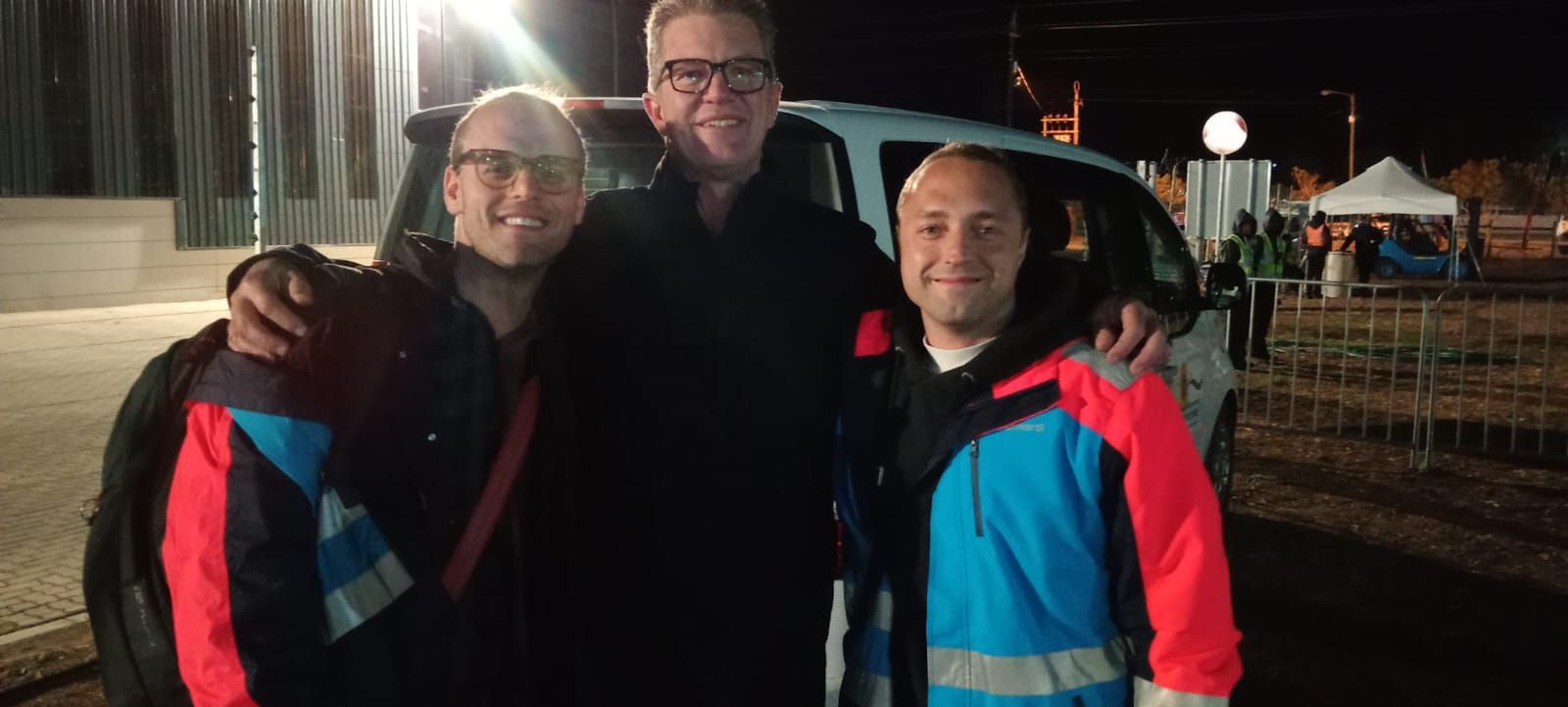 Three people smiling, one with glasses, arms around each other in front of a van at night.