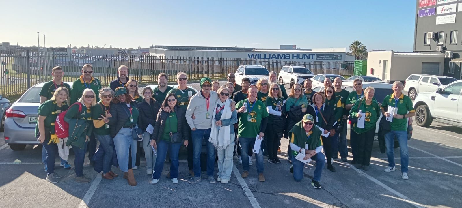 Group of people wearing green shirts posing outdoors in a parking lot. Building and cars in the background.