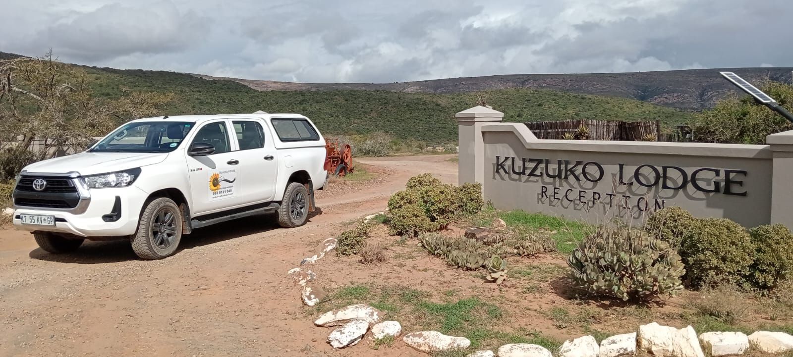 White pickup truck parked near a sign for Kuzuko Lodge Reception. Hills and sky in the background.
