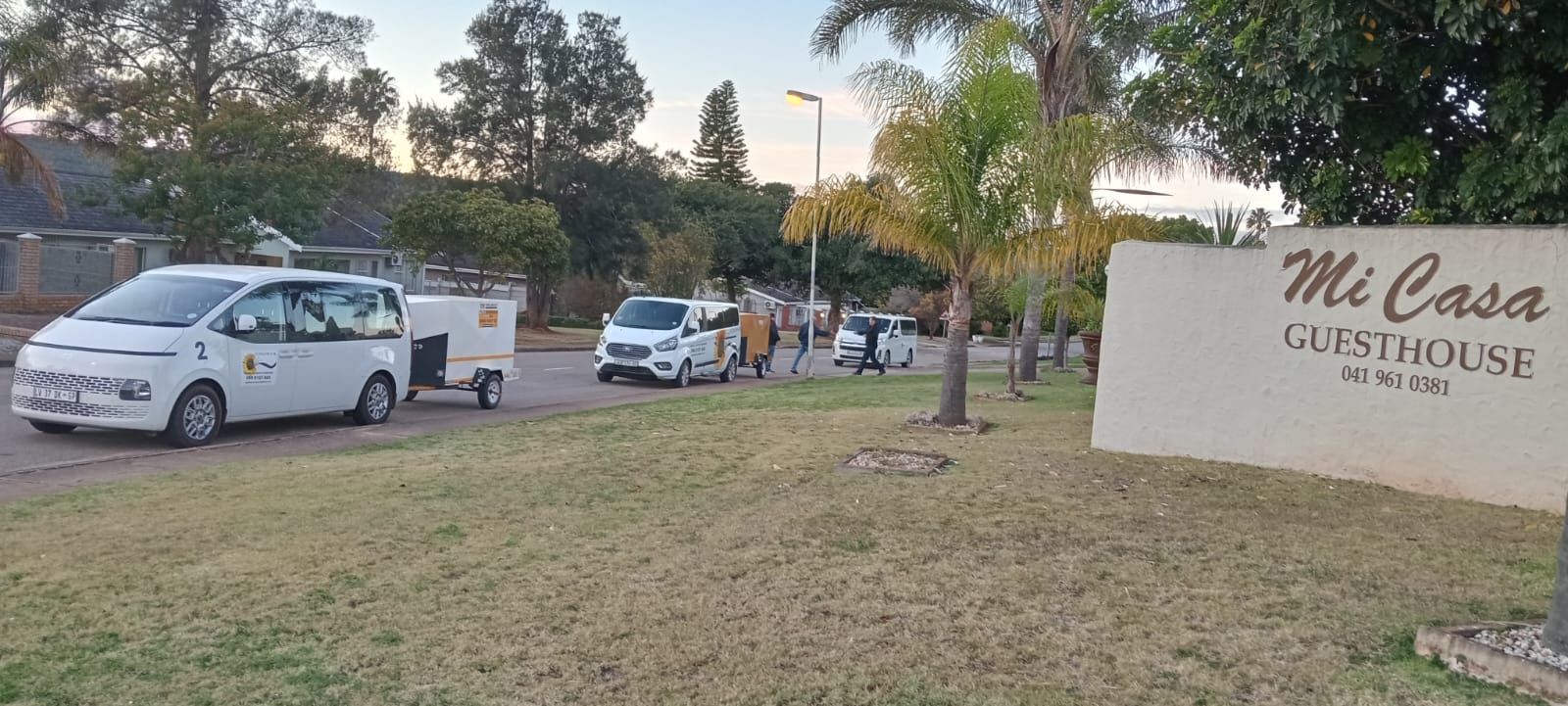 White vans and cars parked in front of Mi Casa Guesthouse on a grassy lawn.