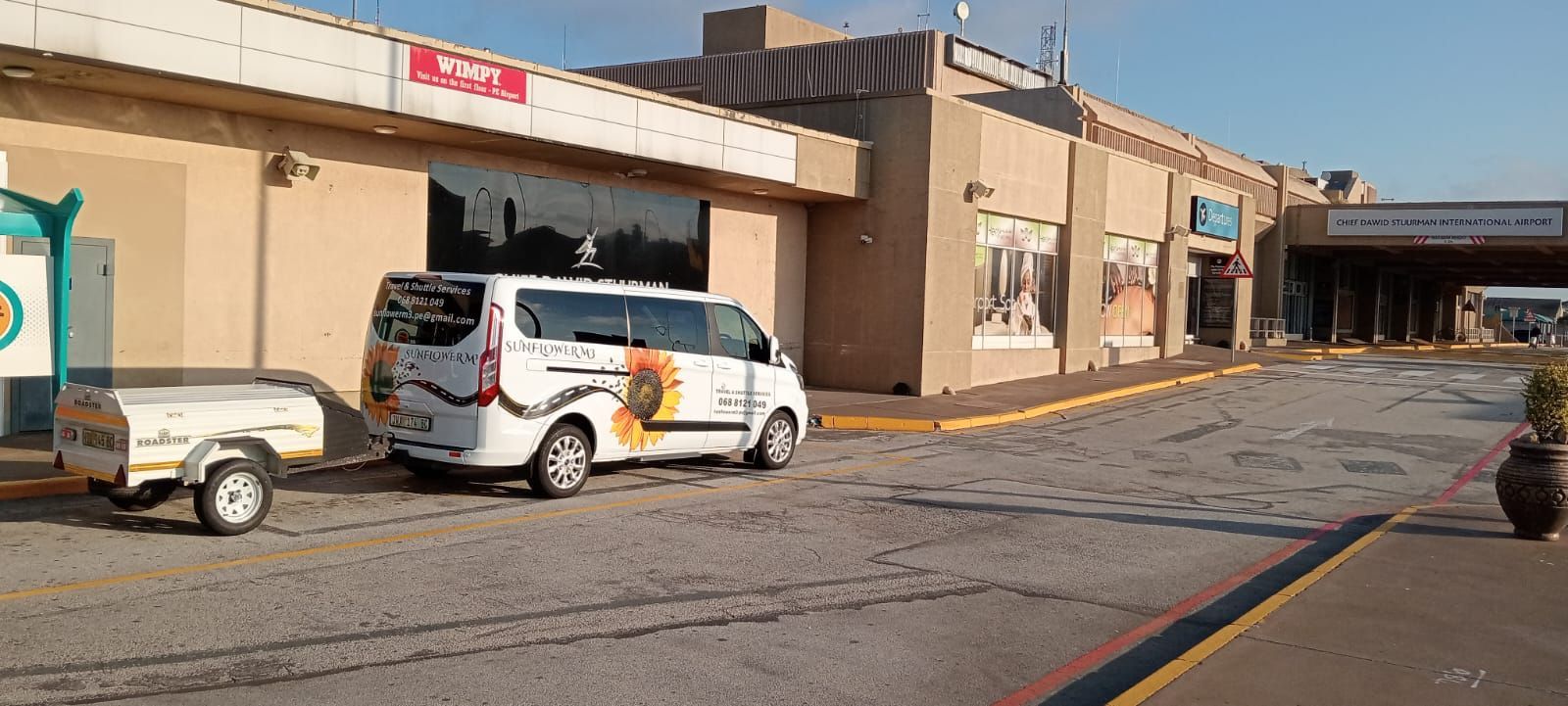 A white van and trailer parked outside a beige building with a glass storefront. The building has a sign that says 