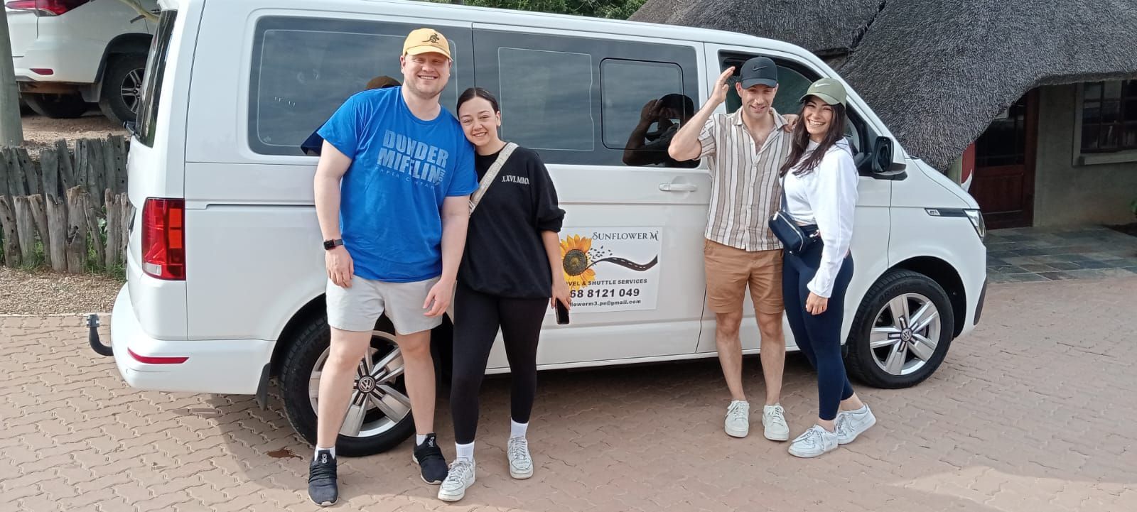 Four people pose by a white van with a safari logo. They stand on sandy ground, near a thatched-roof building.