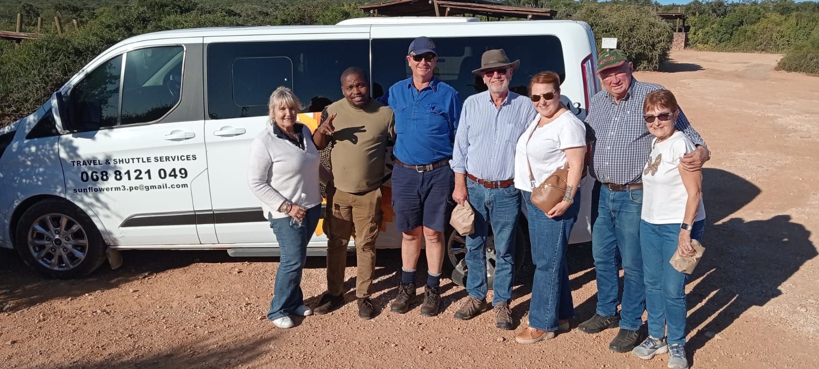 Group of people posing by a white van on a dirt road. Sunny day.