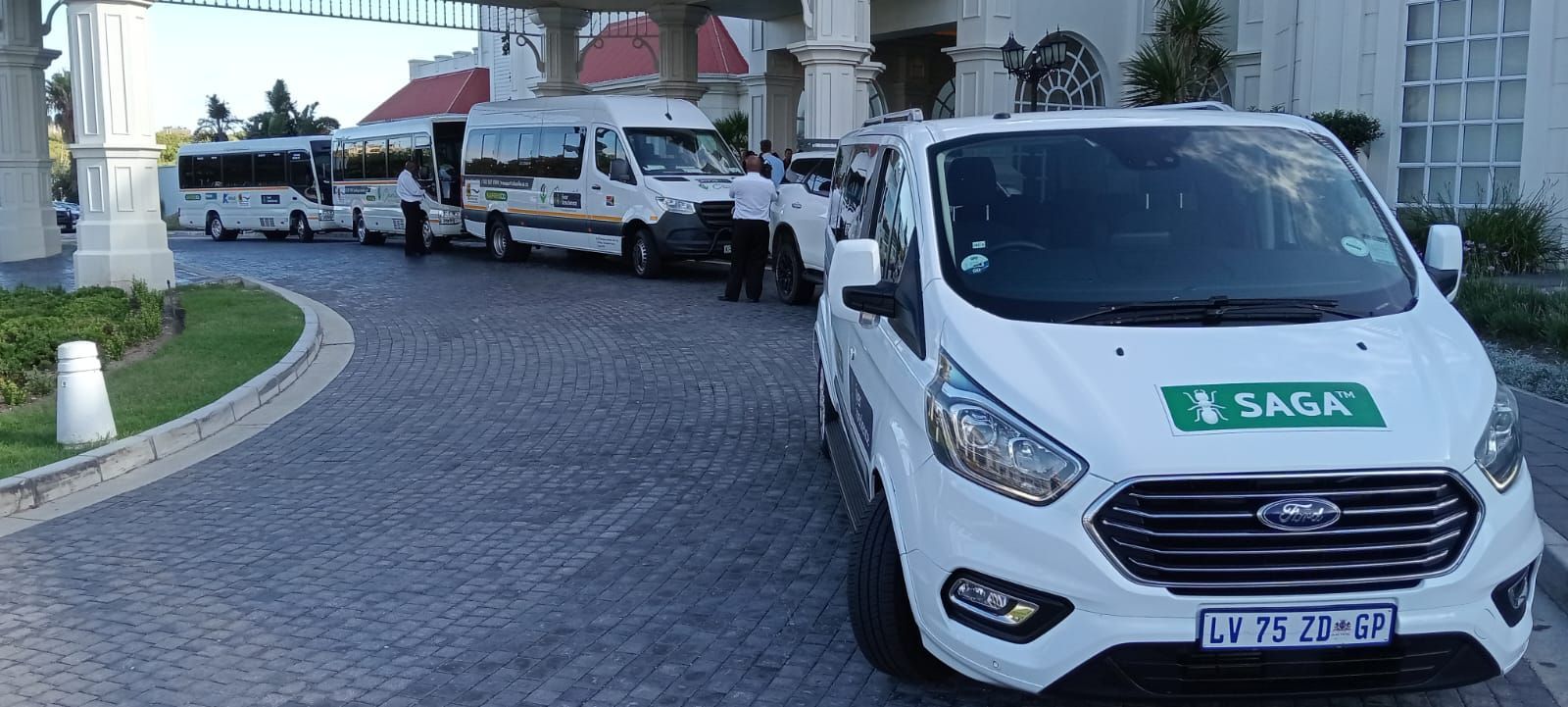 White vans and shuttle buses parked outside a building with pillars. A front van has a green Saga sticker.