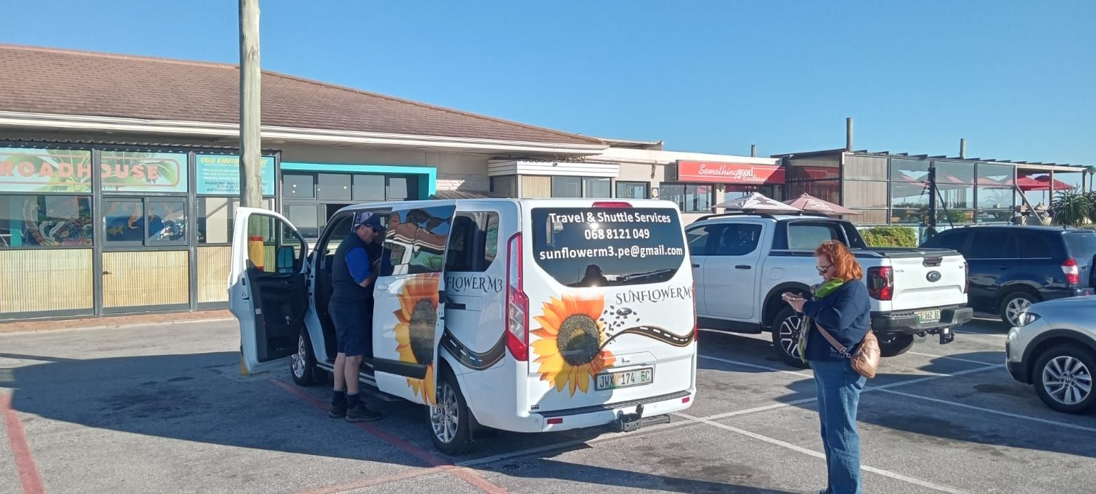 A white van with sunflower decals parked in a lot, doors open. People stand near it. Retail stores in the background.