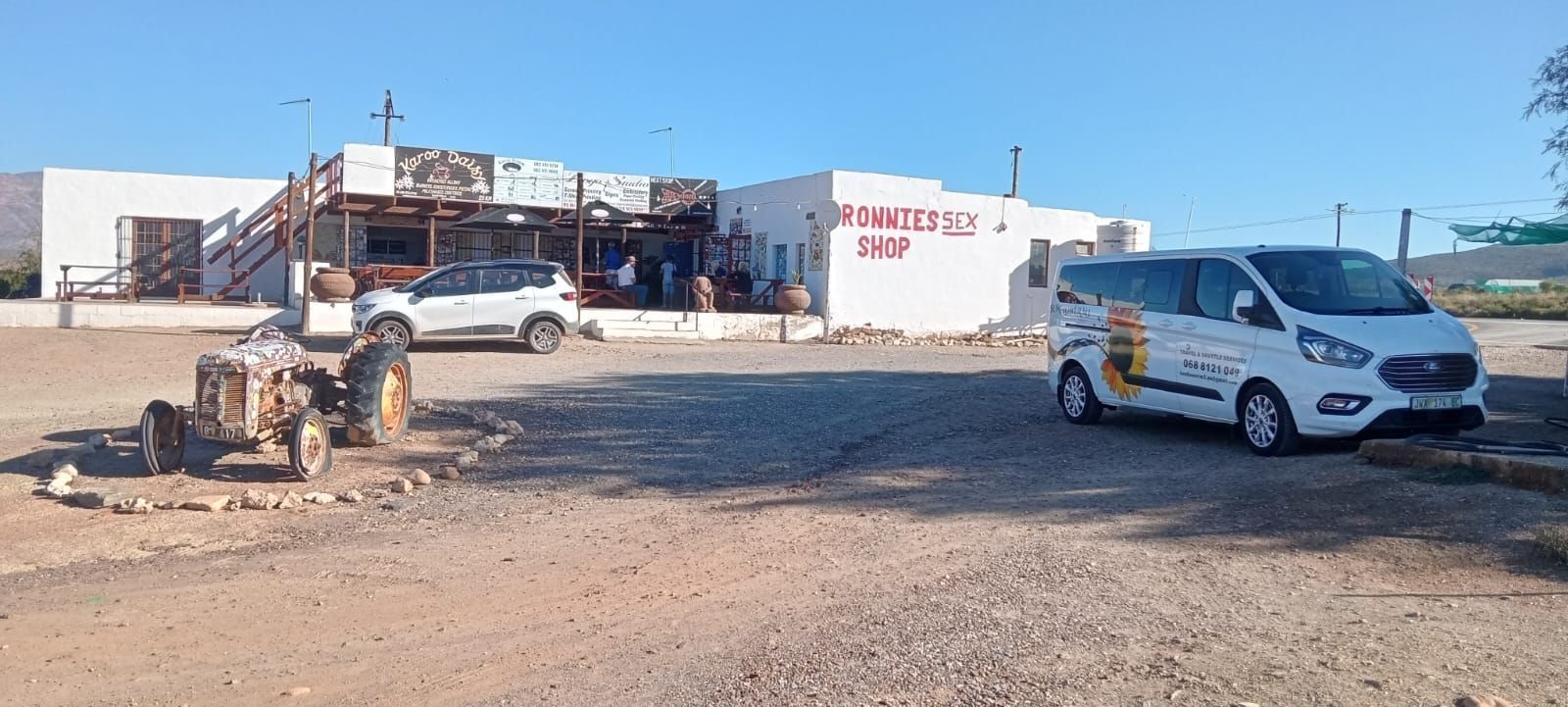A white building with a sign, a white van, and a parked car. Desert landscape.